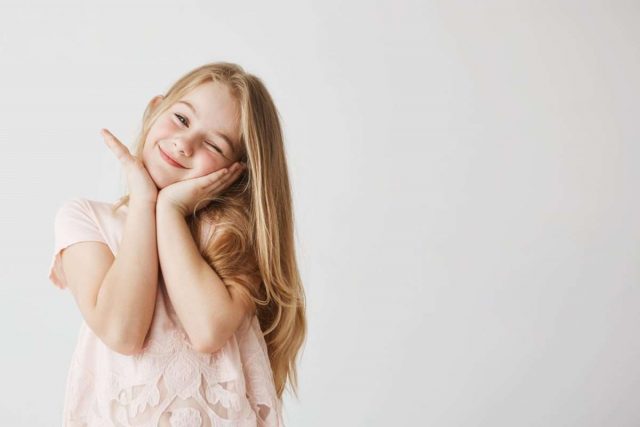 Beutiful little blonde girl smiles at camera winking, posing, touching face with her hands in pink cute dress. Child looking happy and delighted. Copy space.